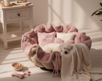 May include: A round, plush, dusty pink pet bed with ruffled edges, containing a sleeping white puppy, a small square pillow with the word Princess, a cream-coloured ribbed pillow, and a draped cream-coloured knit blanket with two small pink toys.