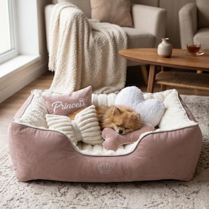 May include: A pink dog bed with a sleeping Pomeranian. The bed has a plush interior and is filled with decorative pillows, including a bow-shaped pillow and a pillow with the word "Princess". A crown design is embroidered on the bed.