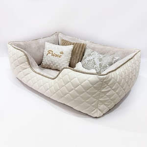 May include: A rectangular pet bed with cream-colored quilted exterior walls and a soft interior lining. It includes three decorative throw pillows: one white textured pillow with the word Prince embroidered in gold, one brown ribbed pillow, and one patterned pillow.