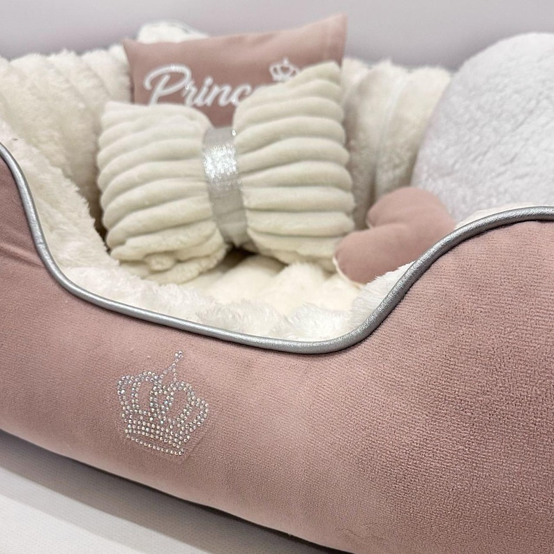 May include: A pink pet bed with a silver trim and a rhinestone crown design. The bed is filled with a fluffy white interior and includes a bow-shaped pillow, a heart-shaped pillow, and a small pillow with the word "Prince" embroidered on it.