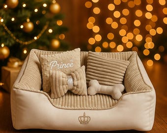 Puede incluir: Una cama para mascotas beige con interior de pana y exterior liso, con una corona dorada bordada en el frente. Dentro hay tres cojines pequeños: uno cuadrado con la palabra Prince, uno rectangular, uno con forma de hueso y un lazo decorativo.