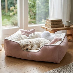May include: A light pink dog bed with a white interior and a sleeping white dog. The bed has a gray trim and includes a pink pillow with the word "Prince" and a crown design. Additional plush toys are also in the bed.