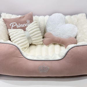 May include: A pink dog bed with a plush interior, featuring a crown design. Includes a bow-shaped pillow with a silver band, a heart-shaped pillow, a bone-shaped toy, and a pillow with the word "Princess".
