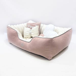 May include: A pink pet bed with a plush, white interior and gray trim. The bed is rectangular with rounded corners and includes several decorative pillows. One pillow has the word "Princess" printed on it.