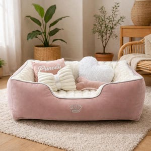 May include: A pink dog bed with a plush interior and a decorative crown emblem. The bed is filled with pillows, including a bow-shaped pillow, a heart-shaped pillow, and a pillow with the word "Princess".