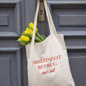 Könnte beinhalten: Eine beigefarbene Tragetasche mit den roten Worten "ARBEITSZEIT BETRUG and chill", die an einem Türgriff hängt. Die Tasche ist mit leuchtend gelben Tulpen gefüllt, die einen Kontrast zur grauen Tür bilden.