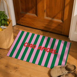 Op de afbeelding: Een rechthoekige deurmat met groene en roze verticale strepen. De woorden "CIAO AMORE" zijn in het rood gedrukt. De mat ligt voor een houten deur, met een geweven tas en sandalen in de buurt.