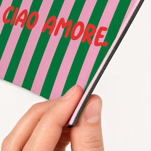 Op de afbeelding: Een rechthoekig item met een roze en groene streepjespatroon en de rode woorden "CIAO AMORE". Het item wordt door een hand vastgehouden, waardoor een zwarte rand zichtbaar wordt.