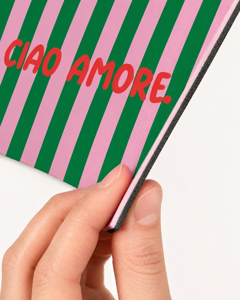 Op de afbeelding: Een rechthoekig item met een roze en groene streepjespatroon en de rode woorden "CIAO AMORE". Het item wordt door een hand vastgehouden, waardoor een zwarte rand zichtbaar wordt.