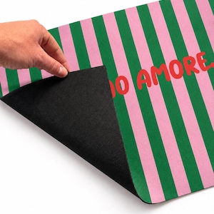 Op de afbeelding: Een deurmat met een zwarte onderkant en een roze en groen gestreept patroon aan de bovenkant. De woorden "IO AMORE" zijn in het rood op de mat gedrukt. De mat wordt opgetild om de zwarte onderkant te onthullen.
