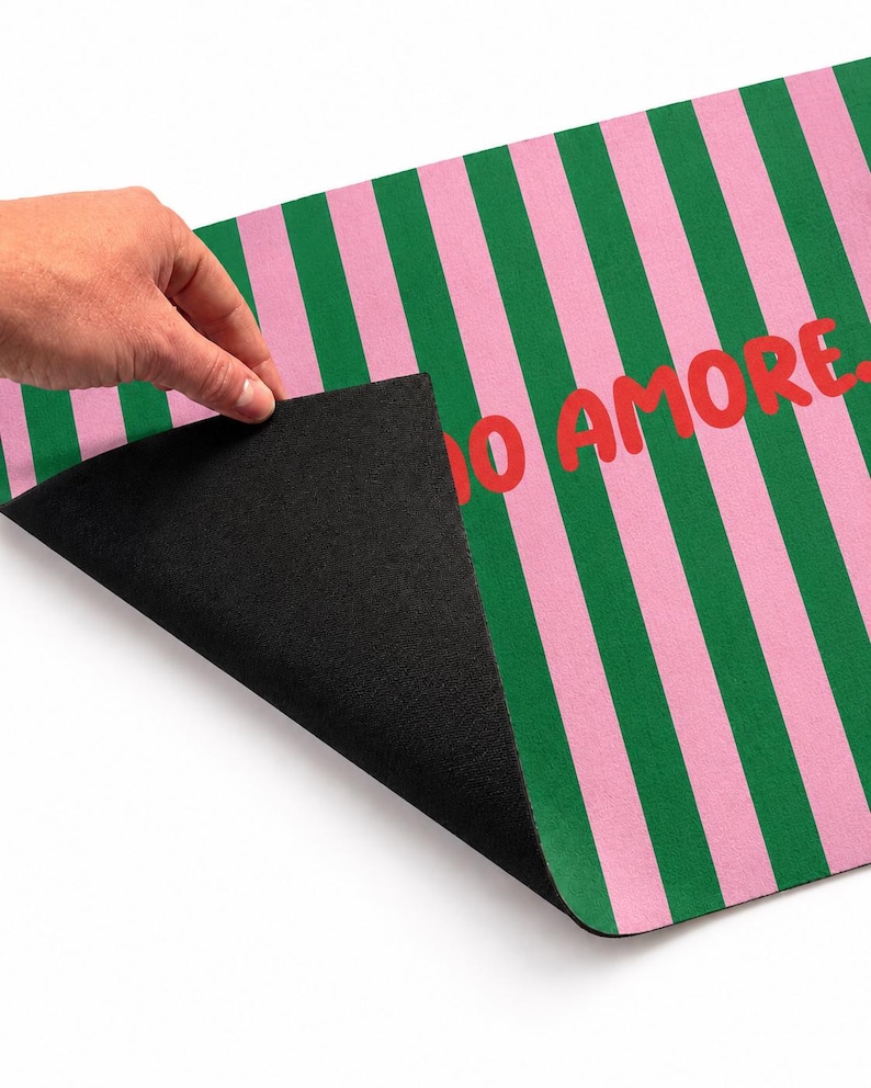 Op de afbeelding: Een deurmat met een zwarte onderkant en een roze en groen gestreept patroon aan de bovenkant. De woorden "IO AMORE" zijn in het rood op de mat gedrukt. De mat wordt opgetild om de zwarte onderkant te onthullen.
