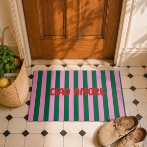 Op de afbeelding: Een rechthoekige deurmat met roze en groene verticale strepen, met de tekst "CIAO AMORE" in het rood. De mat ligt op een betegelde vloer, bij een houten deur en een rieten mand.