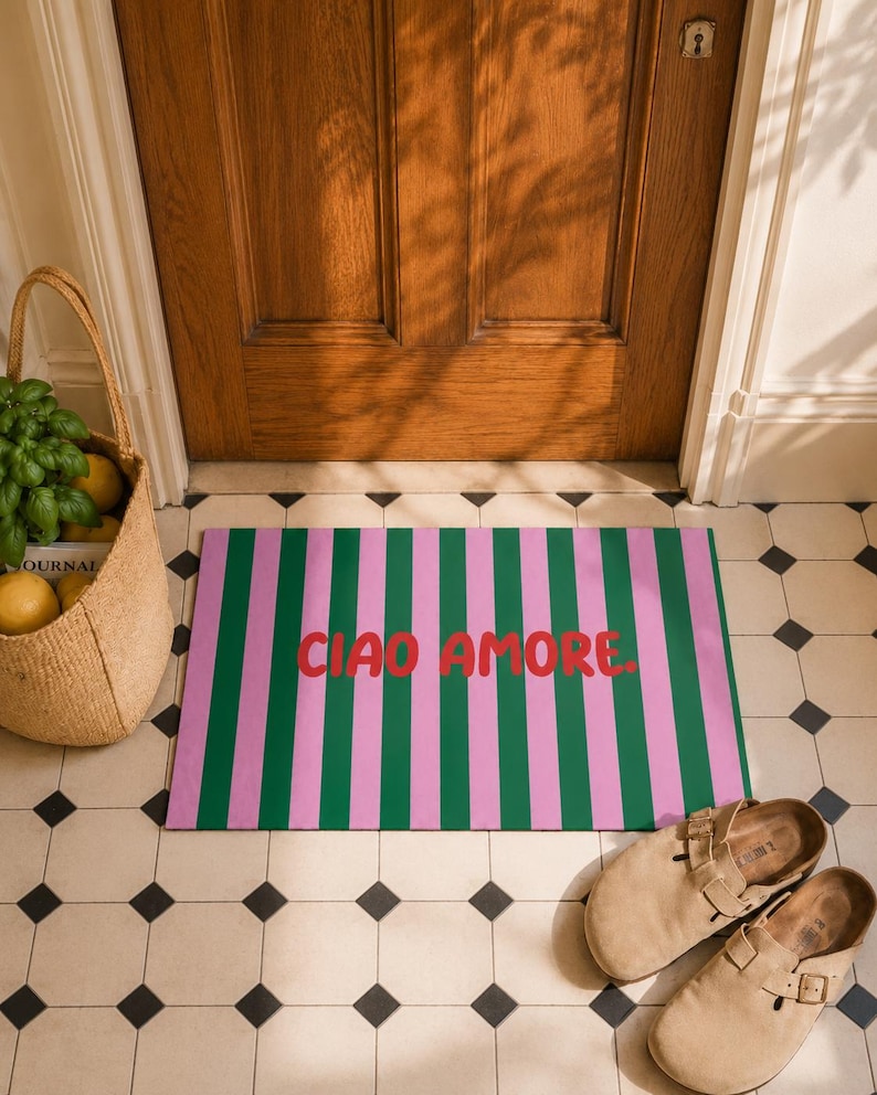 Op de afbeelding: Een rechthoekige deurmat met roze en groene verticale strepen, met de tekst "CIAO AMORE" in het rood. De mat ligt op een betegelde vloer, bij een houten deur en een rieten mand.