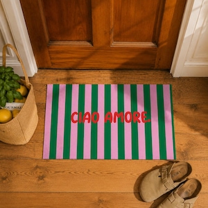 Op de afbeelding: Een rechthoekige deurmat met verticale groene en roze strepen. De woorden "CIAO AMORE" zijn in het rood over het midden gedrukt. De mat ligt voor een houten deur, met een geweven mand en een paar bruinbeige sandalen in de buurt.