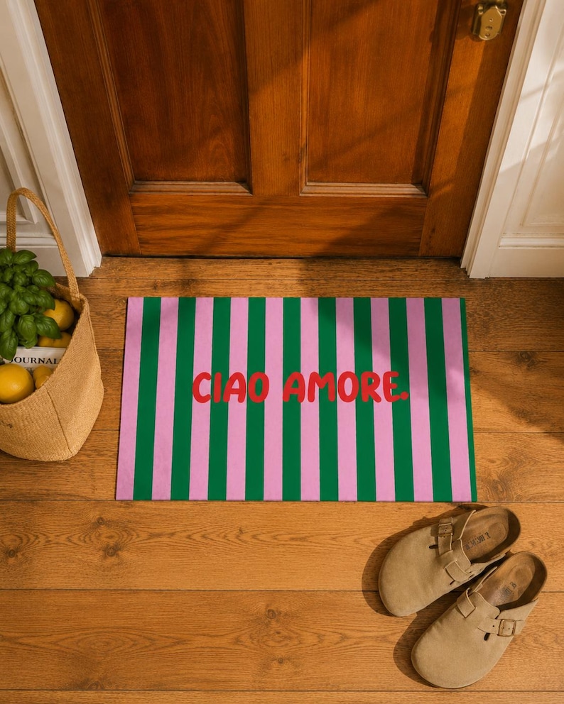 Op de afbeelding: Een rechthoekige deurmat met verticale groene en roze strepen. De woorden "CIAO AMORE" zijn in het rood over het midden gedrukt. De mat ligt voor een houten deur, met een geweven mand en een paar bruinbeige sandalen in de buurt.