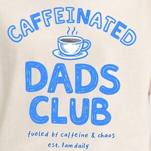 Op de afbeelding: Een crèmekleurig t-shirt met een blauwe afbeelding van een koffiekopje en de tekst "CAFFEINATED DADS CLUB" in grote letters, gevolgd door de kleinere tekst "fueled by caffeine & chaos" en "est. 7am daily" in een eenvoudig blauw lettertype.