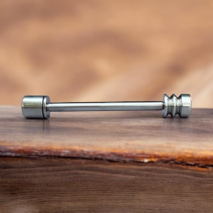 May include: A silver-colored barbell-shaped body jewelry piece. The jewelry has a cylindrical bar with a rounded end on one side and a grooved, rounded end on the other. The piece is displayed on a wooden surface.