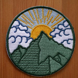 Peut inclure: Écusson brodé circulaire représentant un paysage de montagne. Le motif comprend une chaîne de montagnes vertes, un soleil jaune avec des rayons et des nuages bleus et blancs. Le patch mesure environ 7.6 cm de diamètre. La bordure est verte.