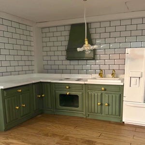May include: A miniature kitchen featuring olive green cabinets, white countertops, and a white tiled backsplash. The kitchen includes a stove, sink, and fridge. A gold-coloured light fixture hangs above the sink. The floor is wooden.