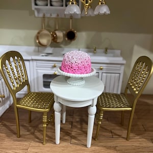 May include: Miniature kitchen scene with a white table and two gold chairs. A pink and white rose-covered cake sits on a white cake stand. The background includes a white cabinet, gold pans, and decorative lighting.