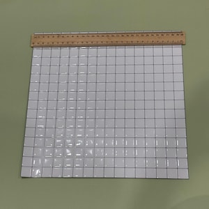 Puede incluir: Una hoja cuadrada de papel cuadriculado blanco con líneas negras finas que forman un patrón uniforme. Una regla de madera está colocada a lo largo del borde superior, mostrando un ancho de aproximadamente 30 cm. La cuadrícula tiene cuadrados.