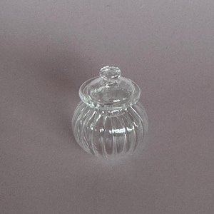 May include: A clear, ribbed glass jar with a matching lid. The jar has a rounded shape and a small knob on top of the lid. The glass is transparent, allowing a view of the contents. The jar is likely used for storing small items.