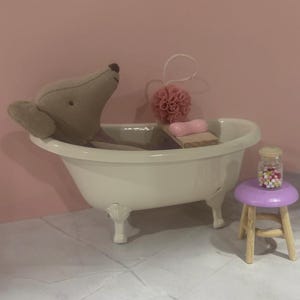 Könnte beinhalten: Eine Miniatur-Badezimmerszene mit einem beigen Stofftier, das in einer weißen Badewanne mit Löwenfüßen entspannt. Ein rosa Badeschwamm, Seife und ein kleiner violetter Hocker mit einem Glas voller bunter Perlen vervollständigen die Szene.