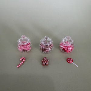 Pode incluir: Três pequenos frascos de vidro transparente cheios de doces listrados em vermelho e branco. Uma bengala de doces, um homem de gengibre e um pirulito em espiral vermelho e branco também estão presentes. Os itens estão dispostos em uma superfície verde clara.