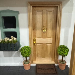 Puede incluir: Miniatura de puerta de entrada de madera con aldaba y manija doradas. Una pequeña alfombra de bienvenida está frente a la puerta. Dos árboles topiarios en macetas flanquean la puerta. Un alféizar con flores blancas está a la izquierda.