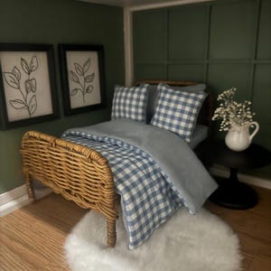 Puede incluir: Escena de dormitorio en miniatura con una cama de mimbre, ropa de cama a cuadros azules y blancos y una alfombra blanca y esponjosa. Dos estampados botánicos enmarcados cuelgan de una pared verde oscuro. Una pequeña mesa auxiliar negra sostiene una jarra blanca con flores.