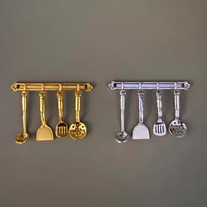 Puede incluir: Dos juegos de utensilios de cocina en miniatura, uno dorado y otro plateado, cada uno colgado de una barra a juego. El juego dorado incluye un cucharón, una espátula, una cuchara ranurada y una espátula ranurada. El juego plateado tiene una disposición similar.