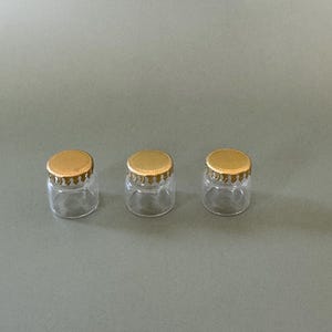 Pode incluir: Três pequenos frascos de vidro transparente com tampas douradas. As tampas têm uma borda decorativa ondulada. Os frascos estão vazios e sobre uma superfície cinza.