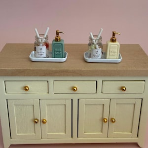 Könnte beinhalten: Zwei Miniatur-Badezimmersets auf einem Holzschrank, jeweils mit einem weißen Tablett, einem durchsichtigen Glasgefäß mit der Aufschrift M IN DOLLHOUSE FLOWER und weißen Stäbchen sowie einem quadratischen Seifenspender mit goldener Pumpe.