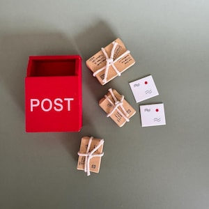 Può includere: Una cassetta delle lettere rossa in miniatura con la scritta "POST" in bianco, accompagnata da piccoli pacchi avvolti in carta marrone e legati con spago bianco. Sono presenti anche due biglietti bianchi con punti rossi e linee ondulate.