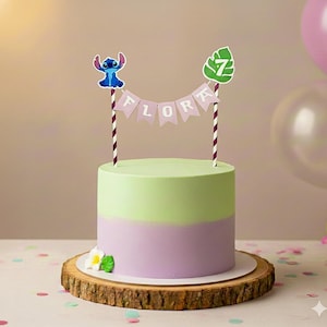 May include: A two-toned cake with green and purple frosting, decorated with a Stitch cake topper and a banner that reads "FLORA". The cake sits on a wooden cake stand and is decorated with a flower.