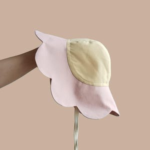 Op de afbeelding: Een lichtroze en gele zonnehoed met een geschulpte rand en een kinband. De hoed wordt tegen een neutrale achtergrond gehouden. De kroon van de hoed is geel en de rand is lichtroze. De kinband is lichtcrème van kleur.