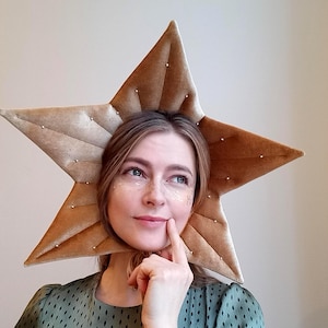 Puede incluir: Un gran tocado dorado en forma de estrella con textura de terciopelo y pequeños tachuelas decorativas. La persona que lo lleva puesto viste un vestido verde con un pequeño patrón repetido. La persona se está tocando la cara.