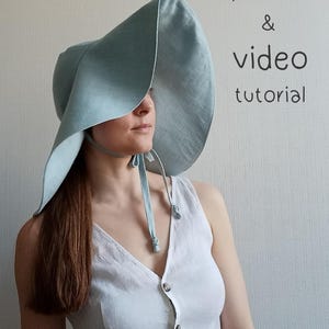 May include: A woman wearing a light blue linen sun hat with a wide brim and a white linen dress. The hat has a tie that is tied under the chin. The text "PDF sewing pattern & video tutorial" is written in black on a white background.
