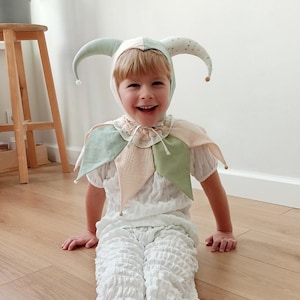 Puede incluir: Un disfraz de buf&oacute;n para ni&ntilde;os en blanco, verde y melocot&oacute;n. El conjunto incluye un gorro de buf&oacute;n con cuernos curvos, un cuello con volantes y pantalones y top a juego. El ni&ntilde;o sonr&iacute;e y est&aacute; sentado en un suelo de madera.
