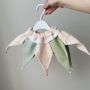 Puede incluir: Un cuello de buf&oacute;n de colores pastel hecho de tela en capas, sostenido por una percha blanca. El cuello presenta secciones alternas de tela rosa y verde, con un borde con volantes y peque&ntilde;os lunares. Las puntas del cuello tienen peque&ntilde;os detalles de bolas de madera y blancas.