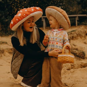 Puede incluir: Dos personas con sombreros en forma de hongo. Uno es rojo con lunares blancos, el otro es marrón. La persona de la derecha sostiene una pequeña cesta marrón claro. La persona de la izquierda lleva un top negro y un chaleco acolchado.