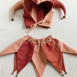 Puede incluir: Un disfraz de buf&oacute;n de dos piezas en tonos rojos y melocot&oacute;n. El conjunto incluye un gorro con capucha con puntas con campanas y un pantal&oacute;n a juego. El gorro tiene un lazo y el pantal&oacute;n tiene detalles de campanas.
