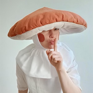 Pode incluir: Um traje de cogumelo caprichoso com uma grande touca cor de ferrugem com uma borda branca pontilhada. O traje inclui uma gola e um vestido brancos, com detalhes vermelhos nas bochechas. A pessoa está pedindo silêncio.