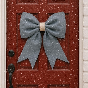 Puede incluir: Un lazo grande de tela gris con un centro tejido está fijado a una puerta roja. El lazo es un adorno decorativo, posiblemente para la temporada navideña. Los copos de nieve caen, lo que se suma al tema invernal. La puerta tiene una manija negra y un marco de ladrillo blanco.