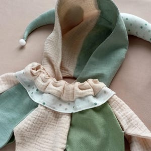 Puede incluir: Un suave gorro de buf&oacute;n en tonos pastel de verde, beige y blanco. El gorro tiene una capucha, puntas largas y curvas y un cuello con volantes con lunares. La tela parece ser un material suave y texturizado, adecuado para un disfraz infantil.