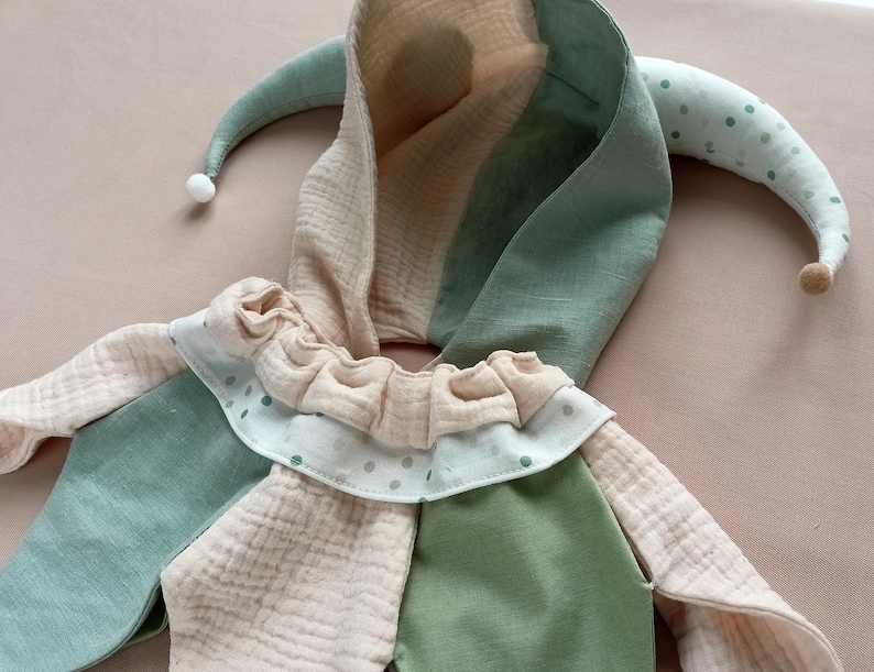 Puede incluir: Un suave gorro de buf&oacute;n en tonos pastel de verde, beige y blanco. El gorro tiene una capucha, puntas largas y curvas y un cuello con volantes con lunares. La tela parece ser un material suave y texturizado, adecuado para un disfraz infantil.