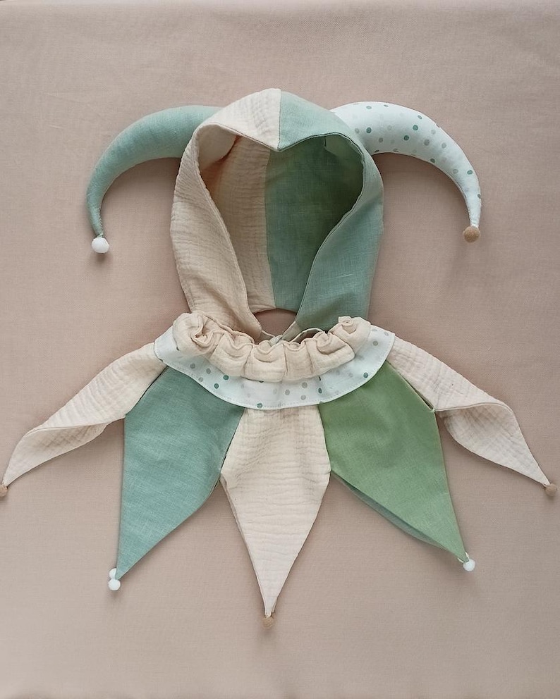 Puede incluir: Una capucha de buf&oacute;n caprichosa en colores pastel suaves. La capucha presenta una combinaci&oacute;n de telas verdes, beige y blancas, con un cuello con volantes y detalles puntiagudos. Tiene dos cuernos largos y curvos con peque&ntilde;os pompones en las puntas, para disfraz o juego.