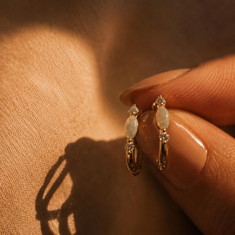 May include: A pair of gold hoop earrings, each featuring a central oval-shaped white opal gemstone flanked by two small, clear round-cut stones. The earrings have a polished metallic finish and are designed as small, circular hoops for the earlobe.