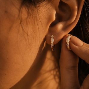 May include: A pair of gold hoop earrings featuring a central oval-shaped opal gemstone flanked by two small, clear, round-cut stones. The earrings are designed with a hinged closure and exhibit a polished metallic finish, reflecting warm light.