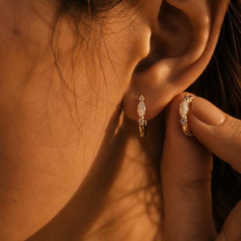 May include: A pair of gold hoop earrings featuring a central oval-shaped opal gemstone flanked by two small, clear, round-cut stones. The earrings are designed with a hinged closure and exhibit a polished metallic finish, reflecting warm light.
